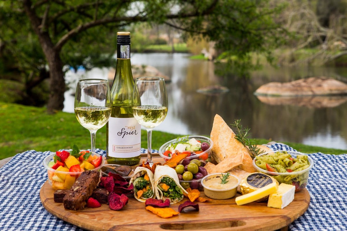 A riverside picnic board with wine, cheese and fruit on a checked blanket